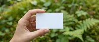 Female hand holding blank white business card outdoors with green nature background, copy space for branding or contact details.