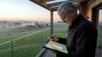 Senior Caucasian man with glasses using tablet on modern balcony overlooking rural farmland at sunset, technology outdoors lifestyle retirement.