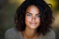 Young mixed race woman with curly dark hair and bright smile wearing casual gray sweater against blurred natural background, close up portrait in warm lighting.
