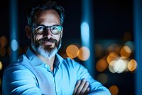 Confident mature Caucasian businessman with gray beard and glasses smiling at night, city lights bokeh background creates professional atmosphere.