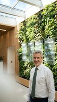Smiling middle-aged Caucasian businessman in green tie standing in modern eco-friendly office with living wall and sustainability data displays.