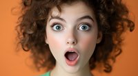 Young woman with curly brown hair and bright blue eyes expressing surprise with open mouth against orange background, close up portrait showing genuine emotion.
