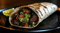 Grilled steak burrito wrapped in flour tortilla with fresh guacamole, diced tomatoes, cilantro and lime garnish served on black plate, close up view of Mexican food.