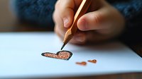 Close-up of hand drawing small orange butterfly with wooden pencil on white paper, artistic process captured in soft natural lighting with shallow depth of field.