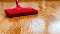 Red microfiber mop cleaning shiny hardwood floor surface, showing contrast between clean and unwiped areas in home interior. Suitable for cleaning service ads.