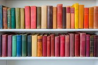 Colorful vintage hardcover books arranged on white wooden shelves, creating rainbow effect with spines in pink, green, yellow, orange, and brown tones. Library collection.