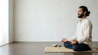 Man with dark hair and beard, South Asian ethnicity, sitting in lotus position on woven mat in minimalist room, meditating or practicing mindfulness. Wearing casual white shirt and jeans. Bright natural light from window. Suitable for wellness, yoga, meditation, mental health, and lifestyle content.