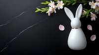 White ceramic bunny figurine with gold collar on black marble surface with pink cherry blossoms and scattered petals creating elegant spring composition.