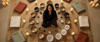 Young woman meditating in center of circular arrangement of singing bowls and cushions for sound healing therapy and wellness practice.