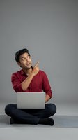 Young man of South Asian descent wearing burgundy shirt, sitting cross-legged with laptop, pointing upward with excited expression. Suitable for tech, education, and business concepts.