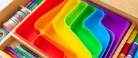 Colorful rainbow plastic organizer trays in wooden drawer with art supplies and colored pencils for creative workspace storage and organization.