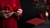 Female hands holding red carnation flowers against dark background with red notebook, creating romantic and elegant mood for Valentine's day or special occasion.
