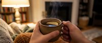 Young adult with medium skin tone holding warm beverage in cozy home setting, wearing knit sweater, evening relaxation and comfort lifestyle moment.