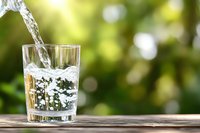 Fresh pure water pouring into clear glass on wooden surface against blurred green natural background with bokeh lights, splash captured in motion.