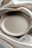Empty ceramic plate with speckled beige glaze on white linen fabric background, creating soft shadows and folds for food photography and table setting styling.