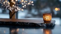 Glowing candle holder on rustic stone surface against blurred winter background with bokeh lights and snowy branches creates cozy atmospheric mood.
