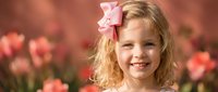 Young blonde girl with pink bow smiling in spring tulip garden. Child portrait with flowers for family photography, greeting cards, seasonal campaigns.