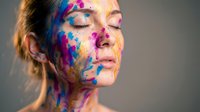 Young woman with eyes closed covered in vibrant colorful paint splatters on face and neck expressing creativity and artistic freedom in studio portrait.