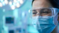Female healthcare professional wearing protective mask and safety glasses in modern medical laboratory with blurred equipment background.