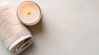 Glowing aromatic candle and rolled white spa towel on light marble surface, creating serene wellness atmosphere for relaxation and meditation practice.