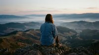 Woman in denim shirt sitting on mountain rock overlooking misty valley landscape at sunrise, contemplative travel and nature exploration scene.