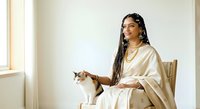 Young woman with dark skin tone and long braided hair wearing a cream-colored shawl, seated in a beige chair while holding and petting a brown and white cat in a bright, minimalist interior setting.