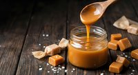 Homemade salted caramel sauce dripping from wooden spoon into glass jar surrounded by caramel candies and sea salt on rustic wooden background for dessert recipes.
