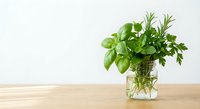 Fresh green herbs basil rosemary parsley in glass jar with water on wooden table against white background for culinary cooking and kitchen decor.