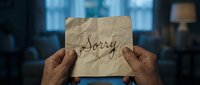 Elderly male hands holding crumpled paper note with handwritten word Sorry in dark moody room, expressing regret and apology concept.