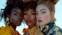 Three diverse fashion models with artistic makeup and statement jewelry pose together against bright colored fabric backdrop, showcasing beauty and inclusivity in modern fashion.