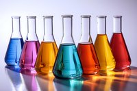 Colorful laboratory glassware containing vibrant liquid solutions in blue, pink, orange, turquoise, red and yellow arranged in row on reflective surface with soft lighting.
