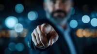Middle-aged businessman in dark suit pointing finger directly at viewer against blurred city lights background, creating powerful confrontational perspective.