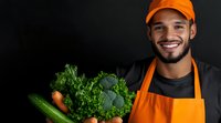Young Hispanic male grocer in orange apron and cap holding fresh organic vegetables including kale, broccoli and carrots against dark background. Friendly smile.