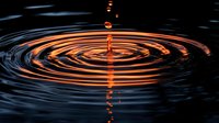 Orange water drop falling into dark liquid creating concentric ripple circles with reflection of light, dramatic macro photography on black background.