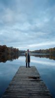 Businessman in coat standing on wooden dock overlooking calm lake with autumn foliage and cloudy sky for contemplation and leadership concepts.