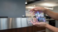 Hands holding transparent tablet with holographic interface displaying digital data and charts in modern office environment for futuristic technology concepts.