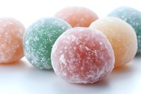 Colorful sugar-coated gummy candy balls in pastel pink, green, and orange on white background, macro shot showing crystallized sugar texture and soft focus.