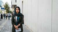Young woman of Middle Eastern descent wearing olive green jacket and teal scarf standing against white wall on urban street, suitable for lifestyle, diversity, and portrait photography.