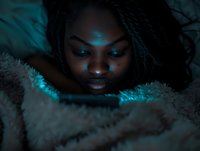 Young African American woman illuminated by blue light from device screen while lying under cozy blanket at night, creating moody atmospheric portrait.