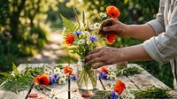 Florist arranging fresh wildflowers in glass vase outdoors, hands holding stems with red poppies and blue cornflowers, garden setting with natural sunlight and blurred green background.