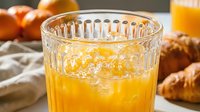 Fresh orange juice with ice in a textured glass, croissants and oranges in the background, bright morning breakfast scene.