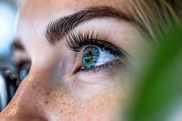 Close-up macro shot of bright blue eye with long eyelashes and natural freckles on light skin, viewed through green leaves creating soft bokeh effect in foreground.