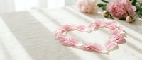 Pink peony petals arranged in a heart shape on a linen surface with soft sunlight and fresh blooms in the background.