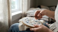 Caucasian woman hands embroidering floral pattern on hoop with scissors near window, cozy home craft hobby, needlework and textile art concept.