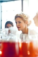 Young Caucasian woman with blonde updo hairstyle and red lipstick smiling during cocktail party gathering, soft focus foreground shows colorful drinks in glasses.