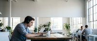 Young professional man working on laptop in modern bright office space with colleagues in background during productive workday.