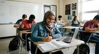 Young African American woman with braids studying in classroom with laptop and notebooks, focused on academic work with diverse students in background.