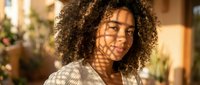 Young African American woman with curly hair smiling warmly in cozy home interior with soft natural lighting and shadow patterns on face.