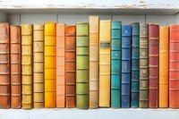 Colorful vintage books arranged in rainbow order on white wooden shelf, showcasing diverse spines with worn edges and faded covers.