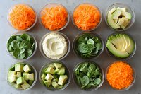 Fresh organic vegetables prepared in glass bowls for healthy meal prep including shredded carrots, diced avocado, leafy greens and chopped ingredients on marble surface.